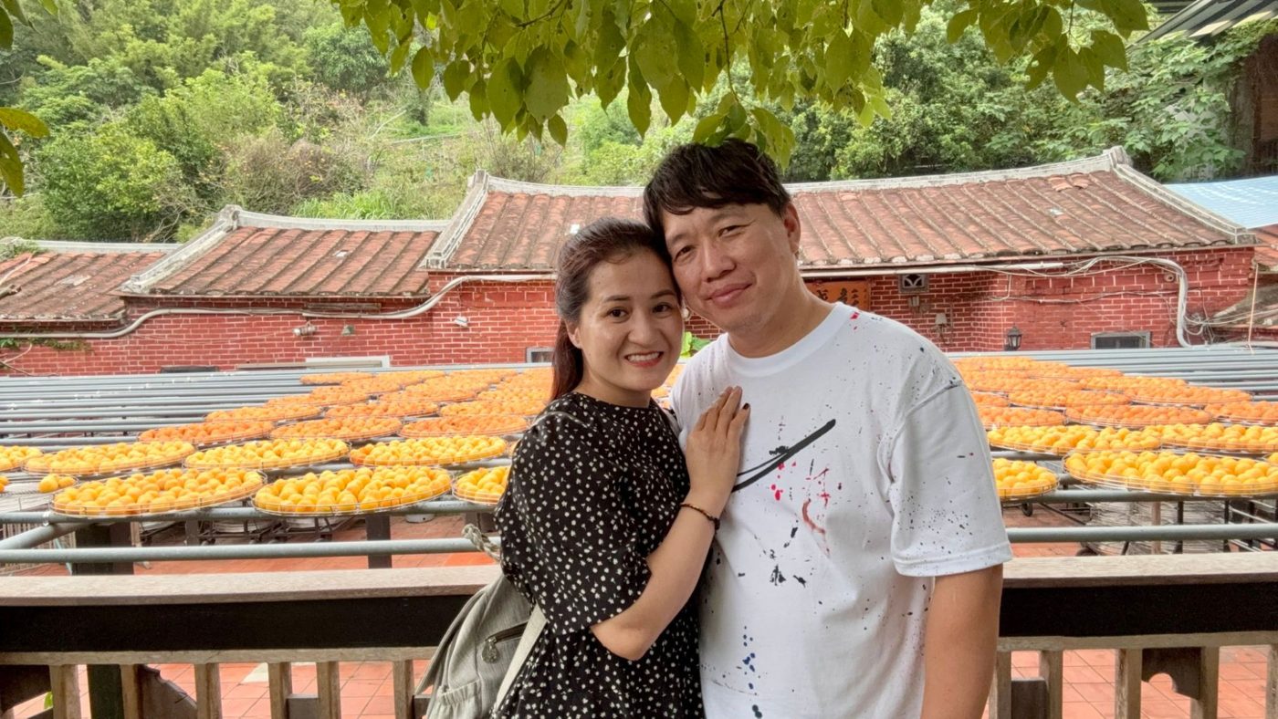 Xiao Cui and her husband smiling in front of trays of drying persimmons and red brick houses